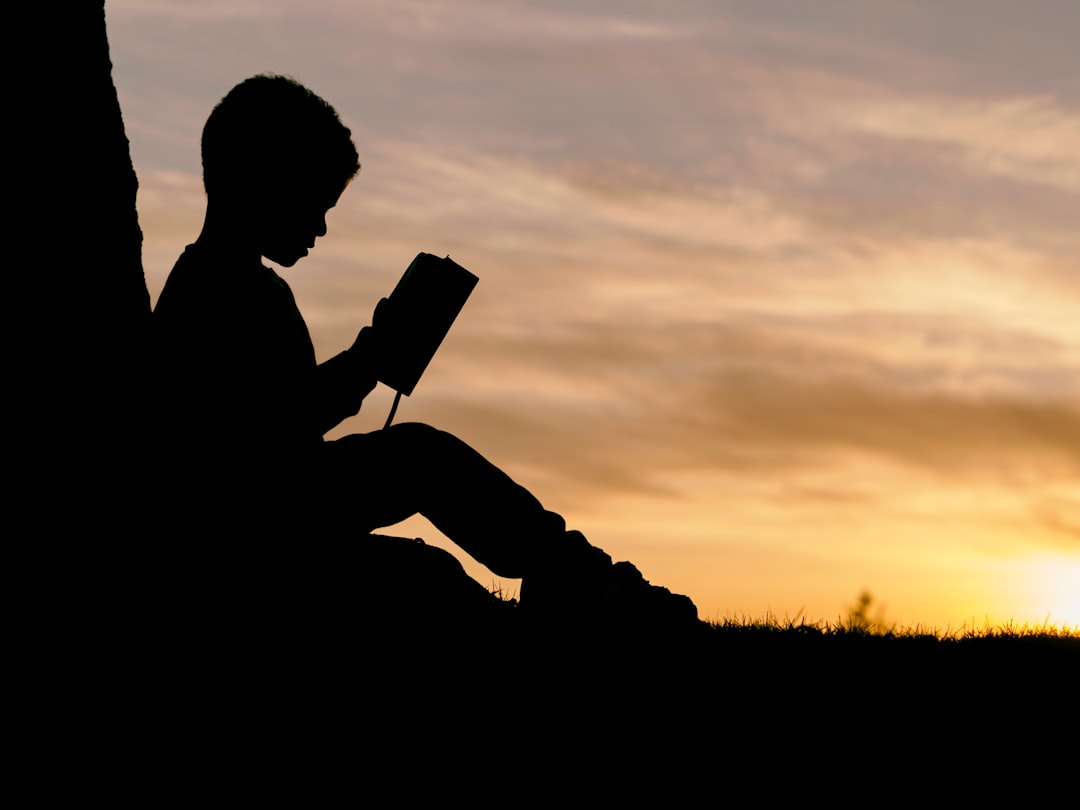 Books(A silhouette of a young child sitting and leaning against a tree trunk while reading a book, set against a bright, warm sunset or sunrise sky.)