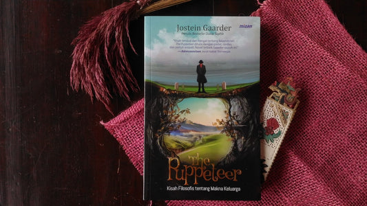 Book titled The Puppeteer by Jostein Gaarder placed on a dark wooden surface with a pink textured cloth, a decorative bookmark, and a dried maroon plant beside it.