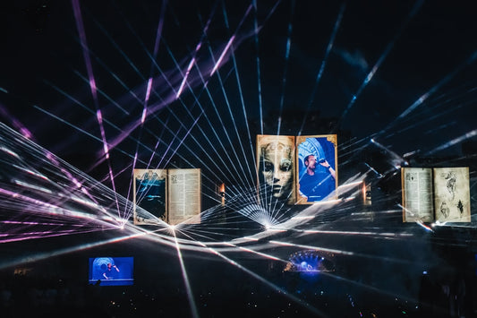 Dramatic shot of a live concert stage with bright pink and purple laser beams crisscrossing the darkness. Large screens display open books and a performer in a blue headpiece