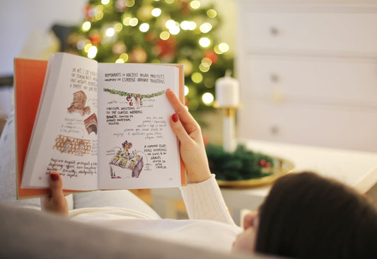 A person relaxing and reading an illustrated book on a couch with a glowing Christmas tree softly blurred in the background.